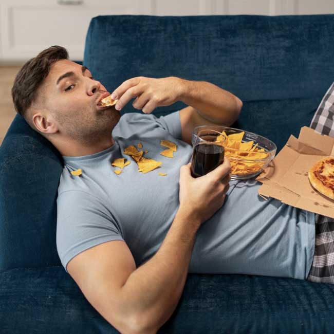 A man lying down on a couch eating chips and drinking soda